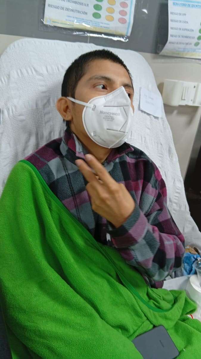 Joseph rapado, con mascarilla y una manta verde, recostado en su cama de hospital