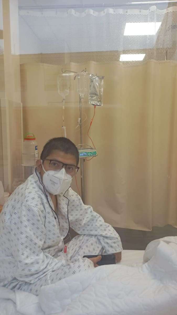 Joseph rapado y con lentes, sentado en su cama de aislamiento detrás de una cortina