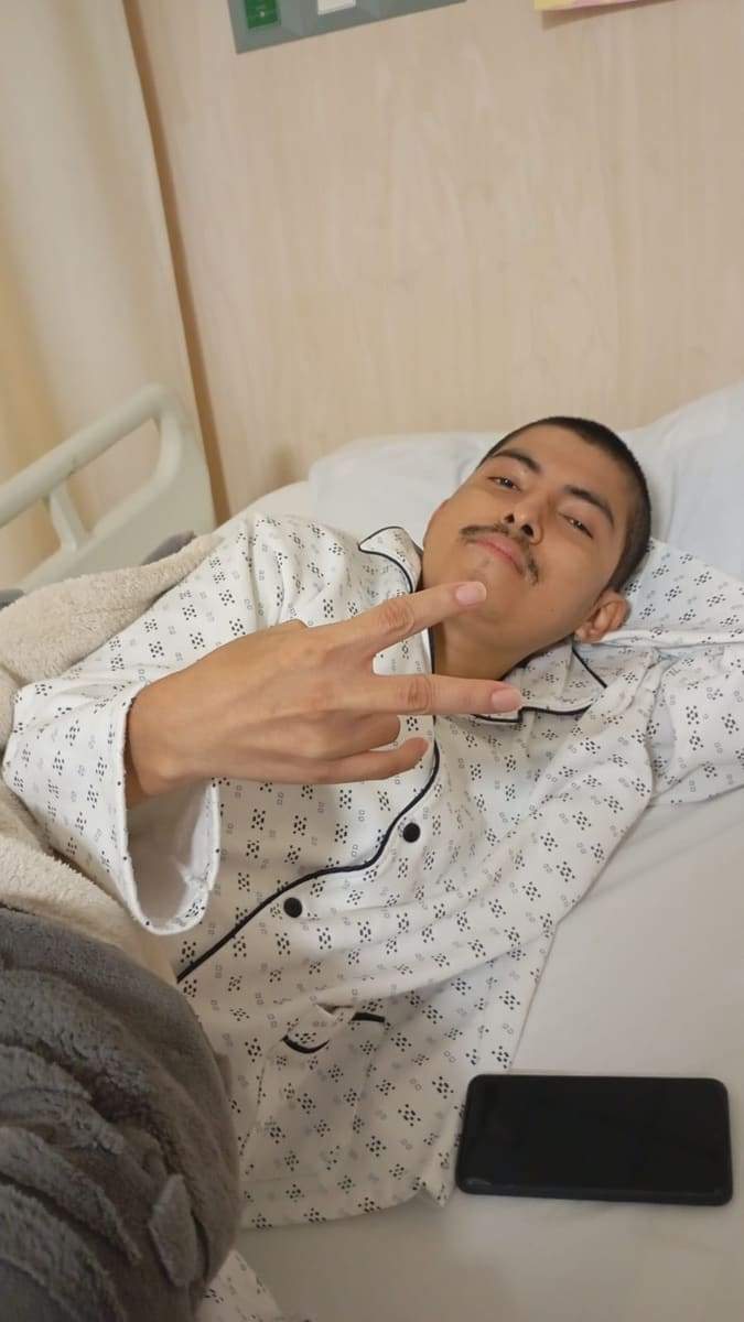 Joseph rapado haciendo el signo de la paz desde su cama de hospital